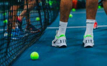 persona jugando tenis con zapatos para jugar tenis marca Nike blancos con verde parado junto a malla de tenis