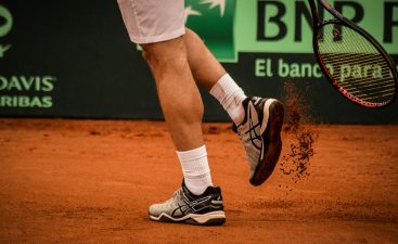 Hombre jugando tenis en cancha de polvo de ladrillo con raqueta de tenis Head usando zapatos para jugar tenis marca Asics.