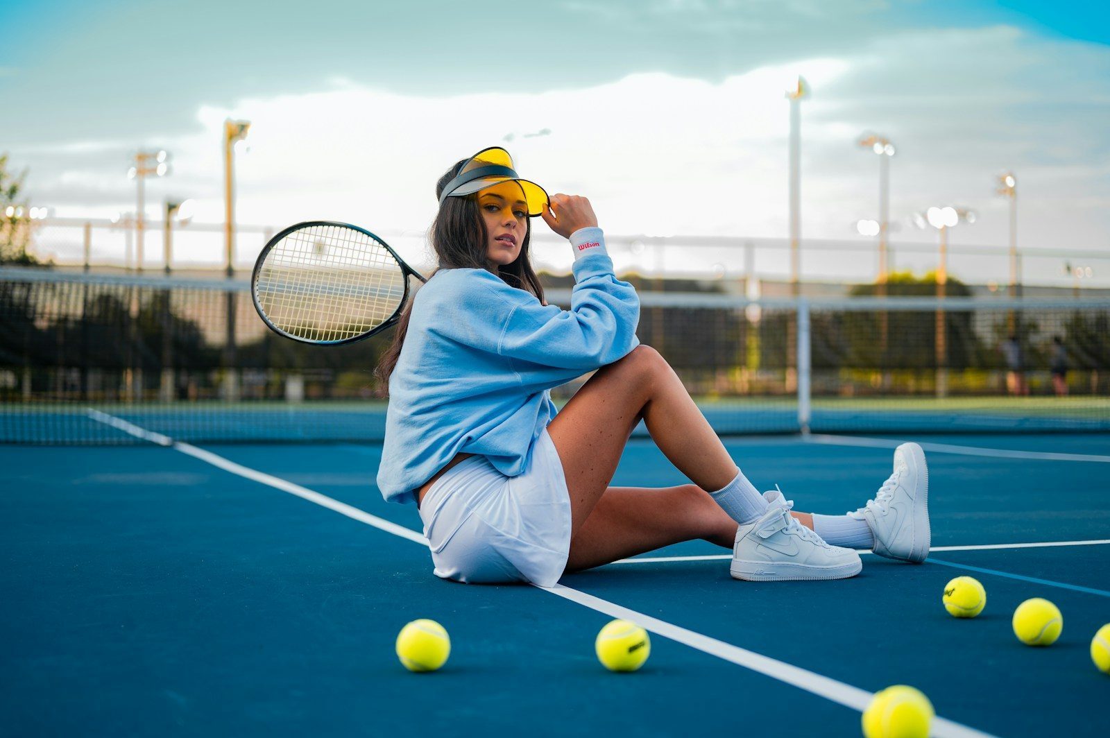 Mujer en una cancha de tenis sintética azul con gorra, falda y raqueta de tenis en mano y sentada en el suelo junto a pelotas de tenis.